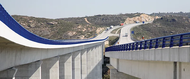 Viaduto em curva com guardas azuis e camiões ao fundo
