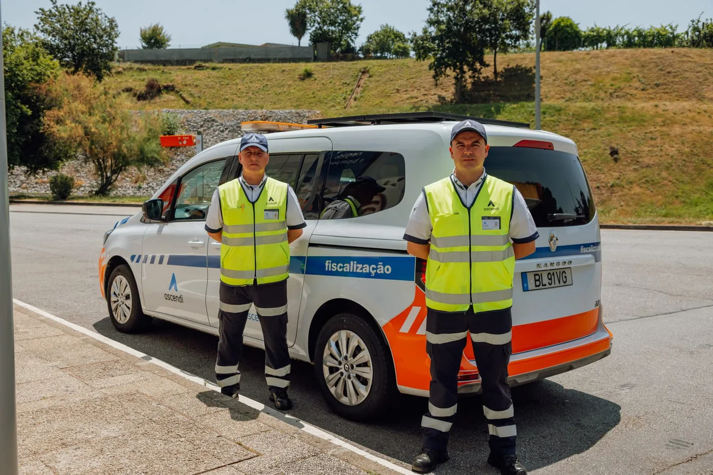 Dois agentes de fiscalização da Ascendi com coletes refletores junto a uma viatura de fiscalização.